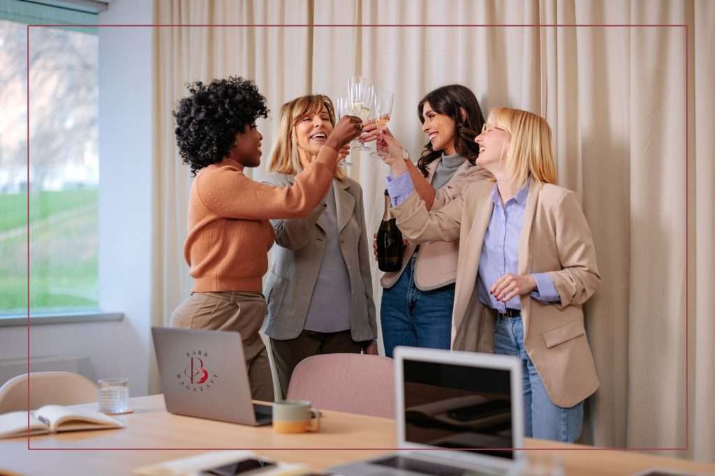 four women standing together and celebrating a successful business collaboration. Laptops are in front of them on a long desk